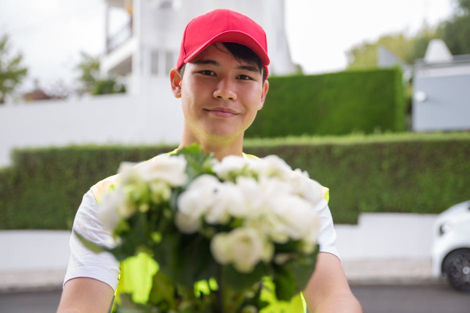 A delivery person wearing a bright pink cap and a high-visibility vest is smiling and handing over a bouquet of fresh, white flowers to a recipient. The bouquet is arranged with white petals and green