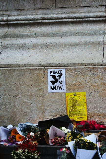 A memorial at the base of a stone building featuring several floral tributes, including bouquets wrapped in white, pink, and purple paper, with flowers such as roses, daisies, and chrysanthemums in va