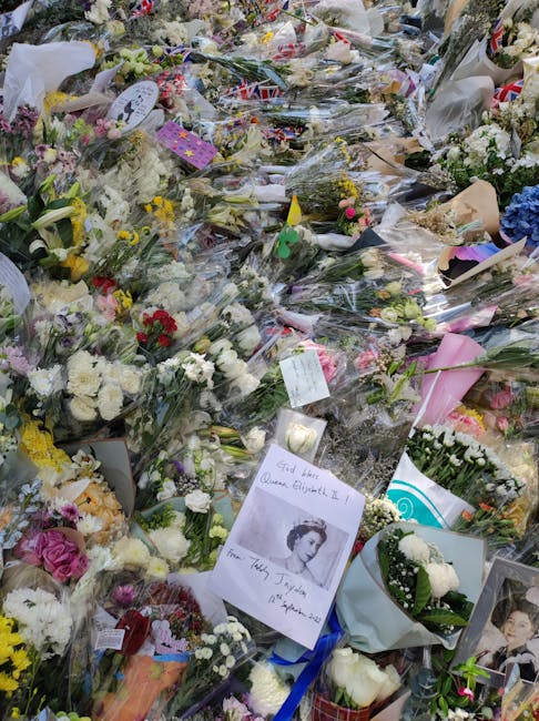 A large collection of floral tributes arranged on the ground, including bouquets of white roses, pink carnations, yellow chrysanthemums, and purple irises, wrapped in clear cellophane and decorative p