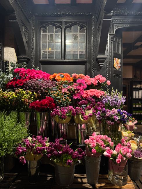 A display of various fresh flowers arranged in metallic vases and containers, including pink, purple, red, orange, and yellow blooms such as carnations, daisies, and other floral varieties. The flower