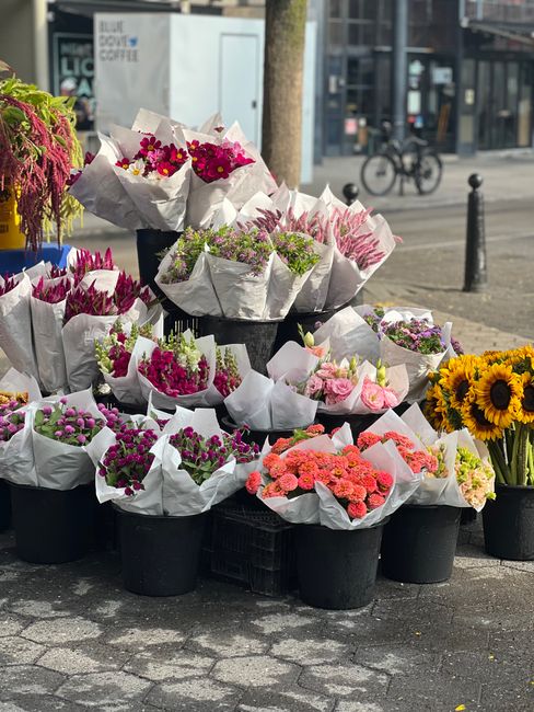 An outdoor display of freshly arranged flower bouquets in black pots, featuring a variety of vibrant blooms such as pink and white roses, bright pink carnations, purple chrysanthemums, and yellow sunf
