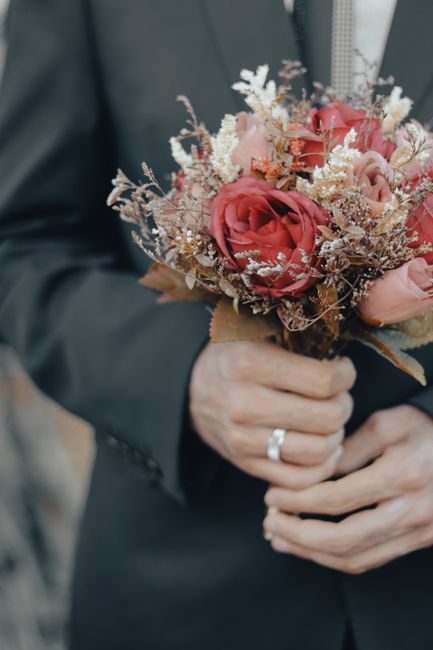 A close-up of a person in a dark suit holding a bouquet of fresh flowers, featuring large pink and coral roses, delicate white and beige fillers, and dried accents arranged in a natural, slightly asym