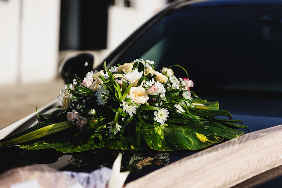 A floral arrangement placed on the hood of a black car, consisting of a bouquet with soft pink and white roses, white daisies, and small white baby's breath, all surrounded by lush green foliage. The 
