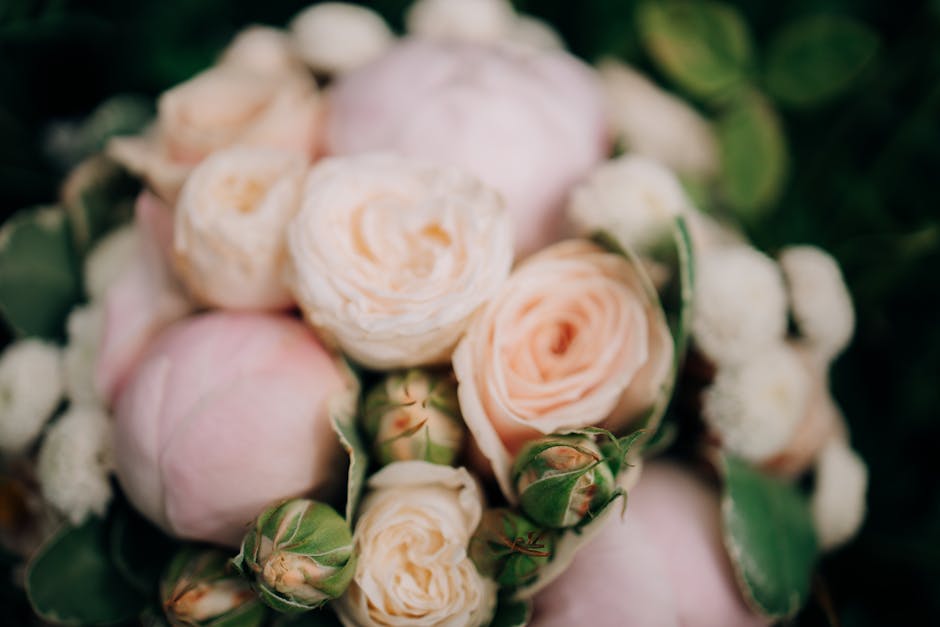 A close-up view of a floral arrangement featuring creamy peach-colored roses with delicate, multi-layered petals that are slightly curled at the edges, complemented by small white buds and surrounded