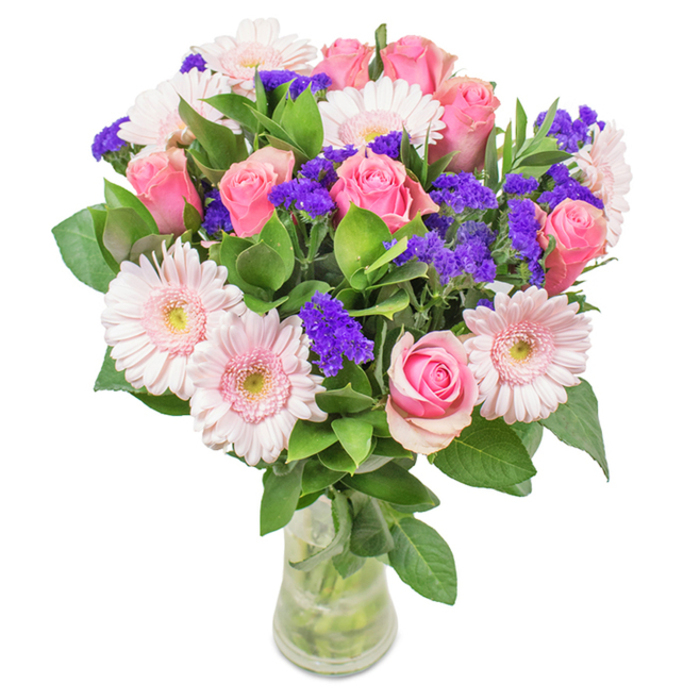 An artfully arranged floral bouquet stands in a crystal-clear glass vase, photographed against a clean, white background. The focal point showcases 7-9 lush pink roses-some petals still curled inward, others just opening into perfect spirals-suggesting both freshness and classic romance. These are complemented by 5-7 pale pink gerbera daisies, their expansive, round heads and nearly white centers evoking springtime walks beside the flower beds of Greenwich Park. Bright purple clusters of statice intersperse the arrangement, adding an energetic counterpoint and tactile richness to the softer forms. The deep green foliage below is abundant and vibrant, calling to mind Greenwich's verdant greenspaces and lending the bouquet a look of local abundance. Clear water shimmers inside the vase, heightening the freshness of the stems as if the bouquet has just arrived to a riverside residence in SE10. Perfect for birthdays, anniversaries, Mother's Day, or thoughtful decoration, this arrangement celebrates joy, love, and Greenwich's garden charm with every detail.